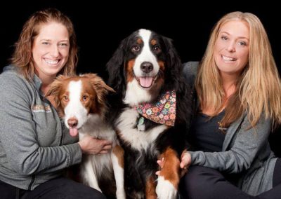 Two smiling women in grey zip-up jackets posing with an Australian Shepherd and a Bernese Mountain Dog wearing a floral bandana